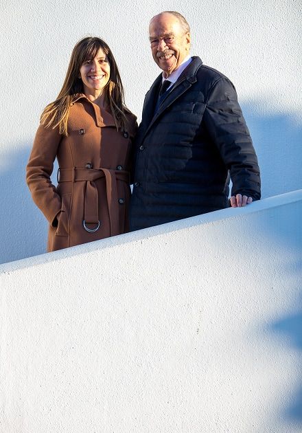 A smiling woman in a brown coat and an older man in a dark jacket stand against a white wall.