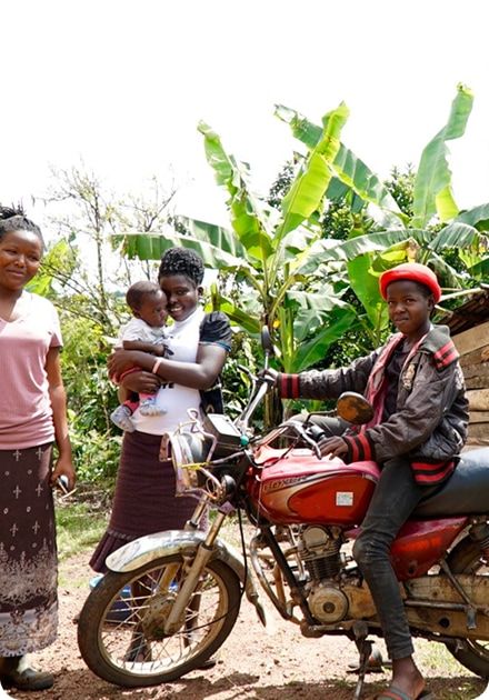 Three people, including a woman holding a baby and a child on a red motorcycle, stand outdoors with banana trees in the background.