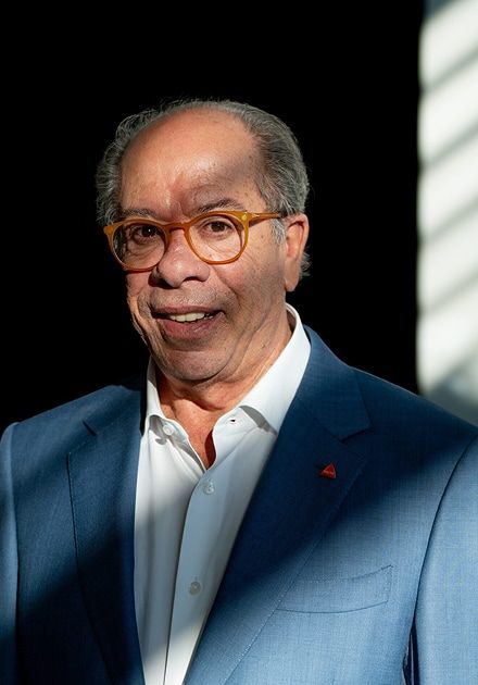 Smiling man with grey hair and orange glasses in a blue suit, lit by strong shadows and light, wearing an orange triangular lapel pin.
