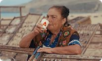 An older woman drinks from a white cup, with fish drying racks and the sea in the background.