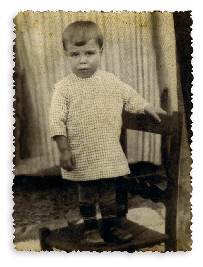 Aged sepia photo of a serious toddler in a checkered shirt standing on a stool next to a chair.