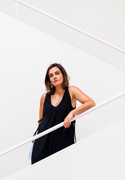 A woman in a dark dress leans on a white staircase railing, looking at the camera.