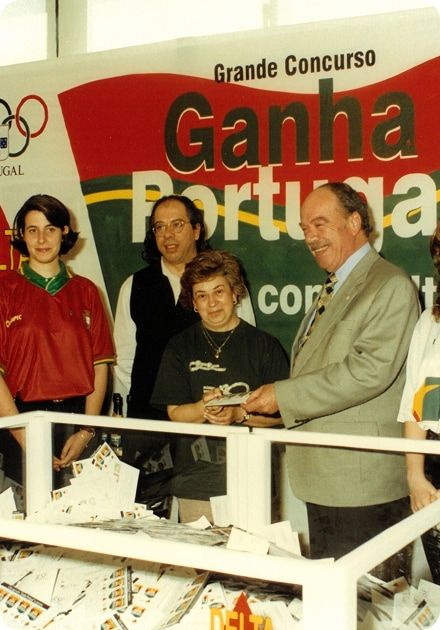 A man in a suit hands a paper to a woman at a "Ganha Portugal" contest, with Olympic rings on a banner and a counter full of entry forms.