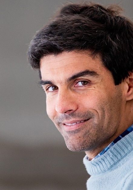 Smiling man with dark hair, light stubble, and brown eyes, wearing a light blue sweater.