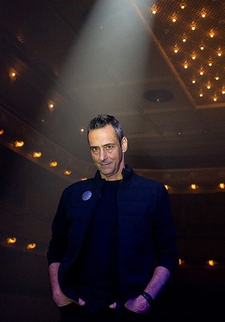 A man with graying hair in a dark vest stands under a spotlight, looking at the camera in a dimly lit venue.