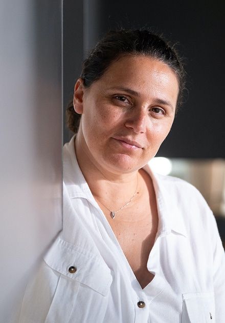 A woman with dark hair in a white shirt leans against a light wall, looking directly at the viewer.