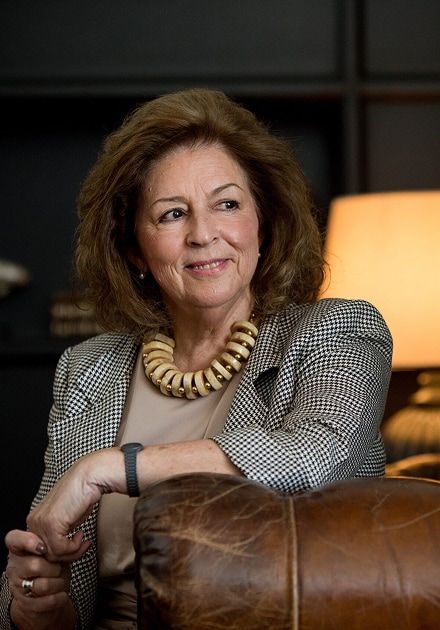 An older woman with brown hair smiles, wearing a houndstooth blazer and a chunky, light-colored necklace, leaning on a leather armchair.
