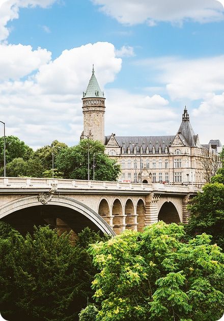 A historic stone arch bridge spans a valley filled with green trees, with a grand building featuring a prominent green-capped tower in the background under a cloudy sky.