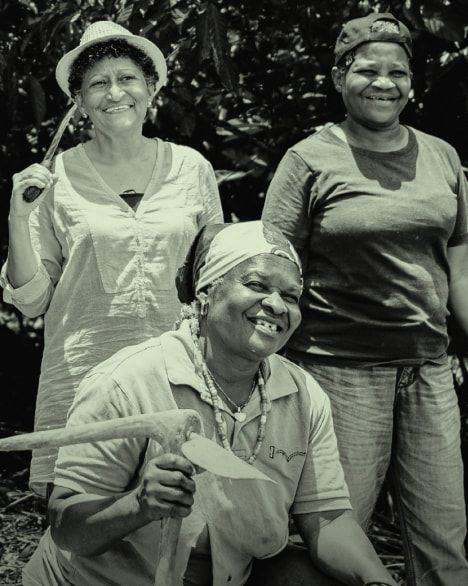 Monochrome portrait of three smiling women, two holding farming tools, against a leafy background.