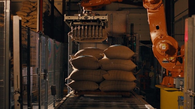 A robotic arm lifts multiple burlap sacks in an automated warehouse.